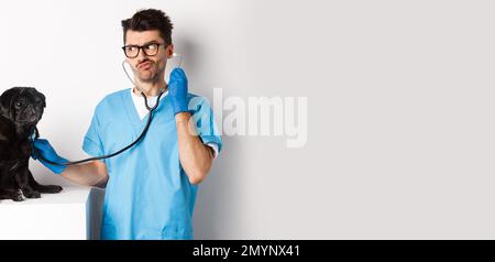 Confused male doctor veterinarian checking dog with stethoscope, looking puzzled, standing over white background Stock Photo