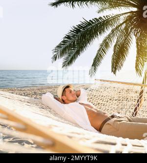 Man relaxing in hammock under green palm leaves on sunlit beach Stock Photo