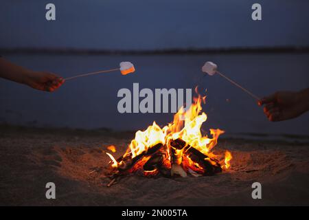 People roasting marshmallows over burning firewood on beach, closeup Stock Photo