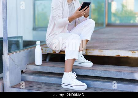 Unrecognizable woman using smartphone sitting on steps near her office. Famela taken a coffee break. Stock Photo