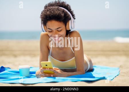 Delighted ethnic female tourist in headphones browsing smartphone on seashore Stock Photo