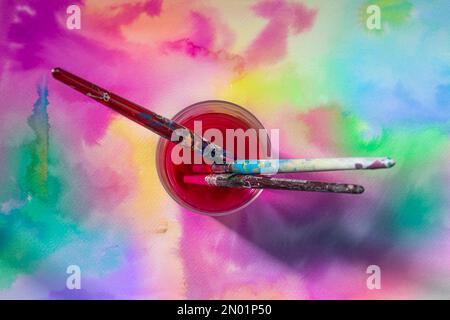 Glass vase with three old paintbrushes with red water on painted paper with watercolors of various colors Stock Photo