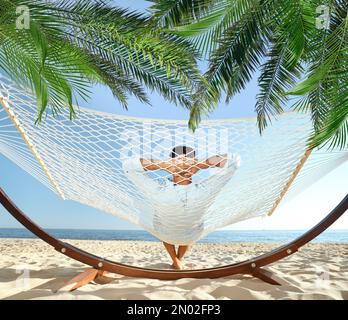 Man relaxing in hammock under green palm leaves on sunlit beach Stock Photo