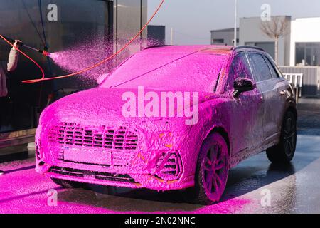 Young woman cleaning her car on self service car wash. Female spread soap using jet sprayer. Pink foam on the car Stock Photo