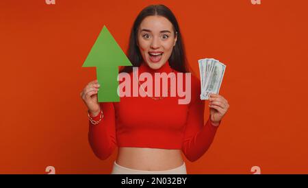 Happy pretty woman winner holding arrow sign pointing up, career growth and money dollar exchange increase. Young adult girl showing up indicating successful development progress on red background Stock Photo