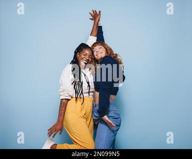 Two best friends dancing and having a good time together in a studio. Happy young women enjoying themselves while standing against a blue background. Stock Photo
