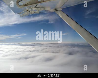 Window view from light general aviation aircraft. Strut and wing of semi-cantilever high-wing airplane flying over the clouds on a sunny day. Stock Photo