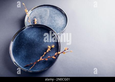 Two handmade round ceramic plates decorated with spring branches. Close up of lacquered blue vintage dishes on grey background. Stylish modern pottery Stock Photo