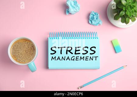 Text Stress Management in notebook on pink background with stationery and cup of coffee, flat lay Stock Photo