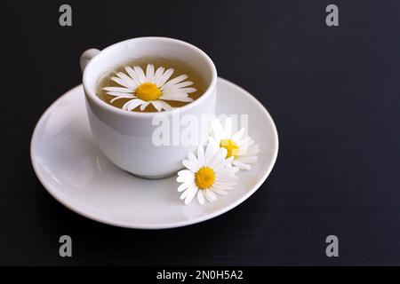 Chamomile tea in a white cup, daisy flowers on saucer. Soothing and healing herbal drink on dark wooden table Stock Photo