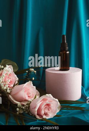 Amber glass dropper bottle on a podium with pink rose flowers. Blue silk curtain background. Skincare products, natural cosmetic. Beauty concept. Vale Stock Photo