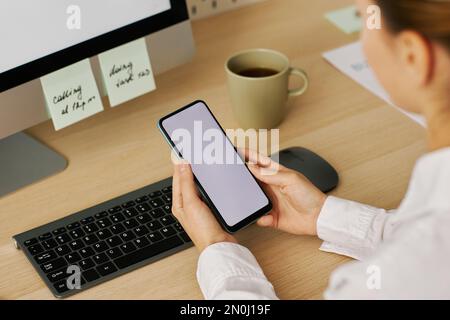 High angle closeup of young woman holding smartphone with white screen mockup over workplace Stock Photo