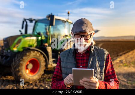 Happy mature farmer working on tablet in front of tractor in field Stock Photo