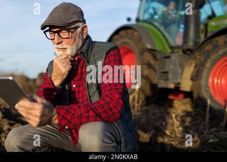 Serious mature farmer working on tablet and thinking about business plans in front of tractor in field Stock Photo