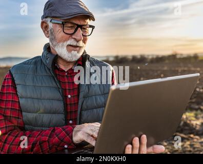 Excited senior farmer smiling and working on laptop in field in autumn time Stock Photo