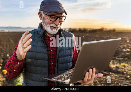 Excited senior farmer smiling and working on laptop in field in autumn time Stock Photo