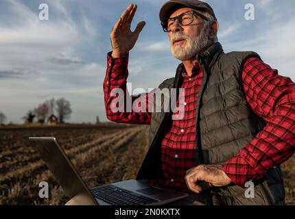 Serious senior farmer working at laptop in field, looking in distance with raised hand Stock Photo