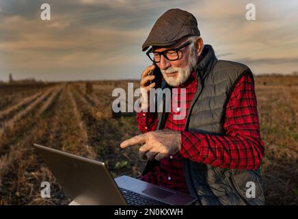 Excited senior farmer talking on phone and looking at laptop in field in autumn time Stock Photo