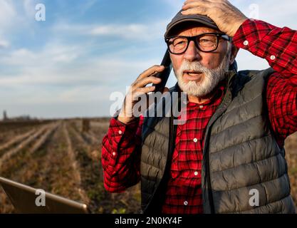 Serious senior farmer talking on phone, holding laptop in lap and holding head in surprise in field Stock Photo