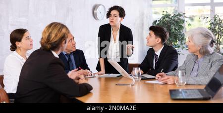 Young Latin female turning on presentation on projector while office coworkers of different ages sitting at table with laptops and notes Stock Photo