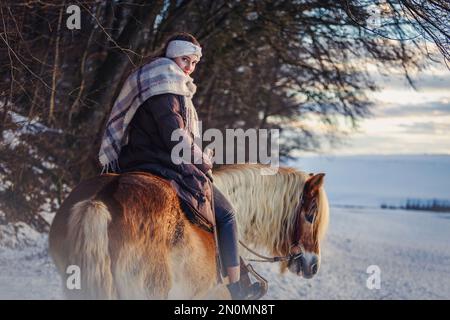 A young equestrian teenage girl rides on her haflinger horse through the snow in the evening during sundown in front of a snowy rural winter landscape Stock Photo