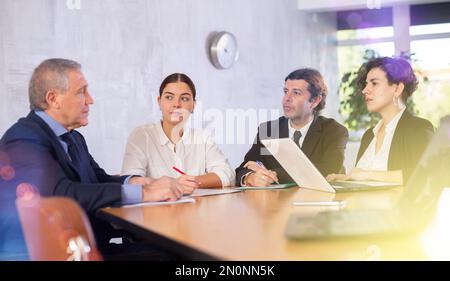 Focused colleagues of different ages sitting around the table and talking before working day in office Stock Photo