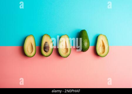 Avocado halves and whole avocado fruit on pink and blue background. Healthy vegan food. Top view, flat lay. Stock Photo