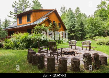 Milled cottage style flat log profile home facade with porch and landscaping plus firepit surrounded by flagstones in summer. Stock Photo