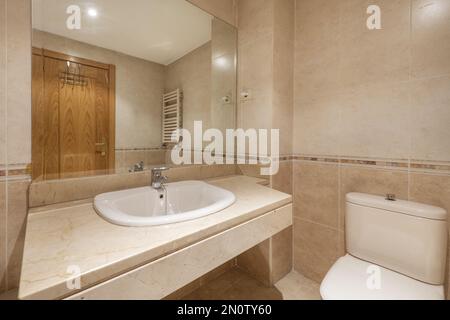 Bathroom with chunky cream marble countertop with white porcelain recessed sink below frameless mirror and cream tiled valanced walls and floors Stock Photo
