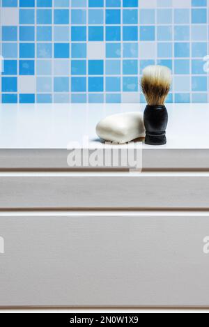 A barber's foam brush next to a bar of white soap in a bathroom with blue tiles and white cabinets Stock Photo