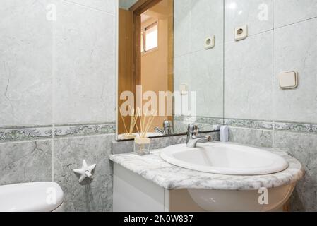 bathroom with a white porcelain sink on a stone countertop below a frameless mirror attached to the wall and valance tiles Stock Photo