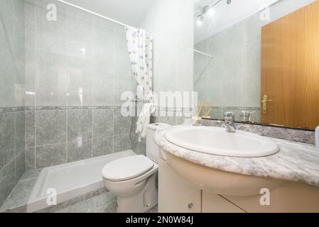 bathroom with white porcelain sink on stone countertop below frameless mirror attached to wall, walk-in shower with curtains and valance tile Stock Photo