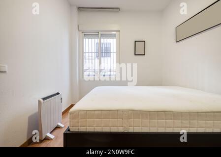 Bedroom mattress without bedding, an electric radiator and window with metal bars Stock Photo
