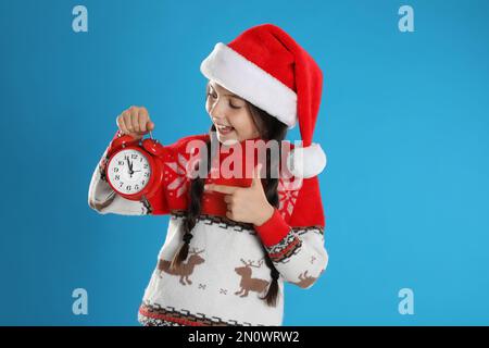 Girl in Santa hat with alarm clock on light blue background. New Year countdown Stock Photo