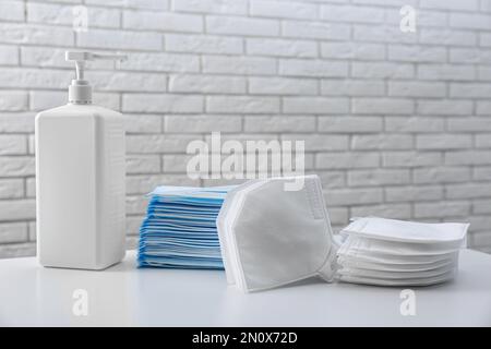 Hand sanitizer and respiratory masks on table near white brick wall. Protective essentials during COVID-19 pandemic Stock Photo