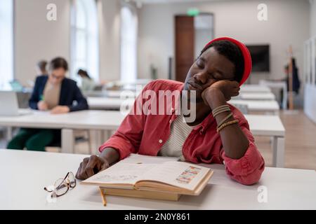 Sleepy lazy African American man falls asleep sits in library with head resting on desk with books Stock Photo