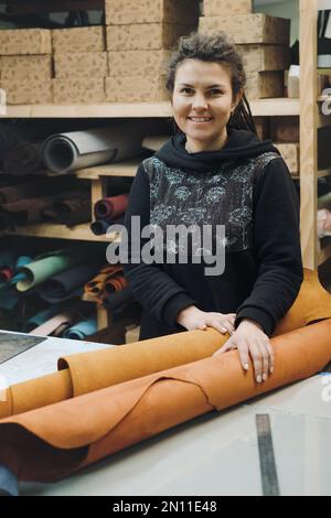 Tailor woman with fabric rolls. Pretty brunette designer looking camera while holding pile of leather rolls in workshop. Occupation concept. Small Stock Photo