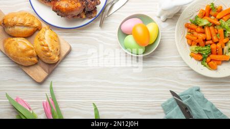 Easter food on white rustic table: pastel colored eggs, roasted chicken and vegetables, buns and spring flowers tulips top view flay lay, Easter famil Stock Photo