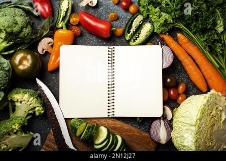 Empty paper notepad and assorted fresh vegetables on rustic concrete table, wooden cutting board, kitchen knife, cooking menu planning, diet and veget Stock Photo