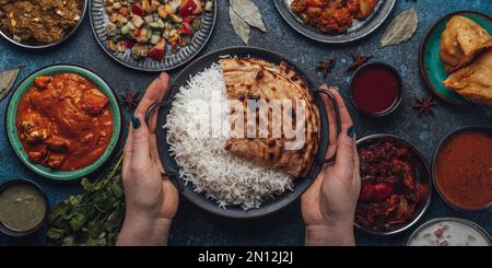 Assorted Indian ethnic food buffet on rustic concrete table from above: curry, fried samosa, rice biryani, dal, paneer, chapatti, naan, chicken tikka Stock Photo