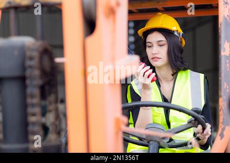 Young asian woman is foreman using radio for communication while driving forklift at distribution warehouse, engineer working and shipping container a Stock Photo