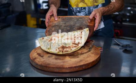 A cooked hot tortilla is cut into pieces and put down on a wooden plate by the chef in the restaurant kitchen. Stock Photo