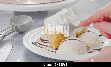 Slice of a freshly baked apple cake with whipped cream topping, served with vanilla ice cream and chocolate close-up on a white plate Stock Photo