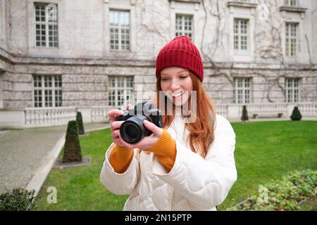 Happy redhead girl tourist, takes photos, photographer with professional camera walks around city and captures beautiful pictures. Stock Photo