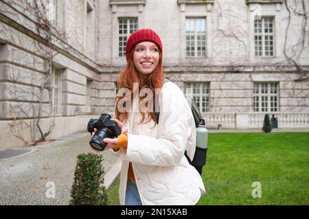 Happy redhead girl tourist, takes photos, photographer with professional camera walks around city and captures beautiful pictures. Stock Photo