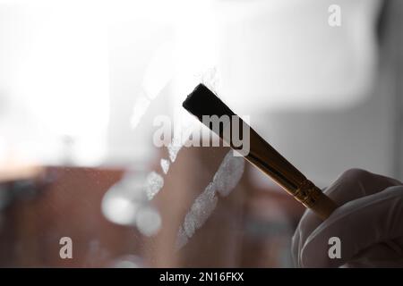 Detective using brush and powder to reveal fingerprints on glass surface indoors, closeup Stock Photo