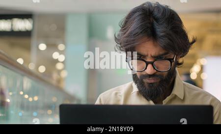 Focused adult ethnic Arabian Indian businessman in glasses typing on computer working indoors. Serious male manager man executive work with laptop Stock Photo