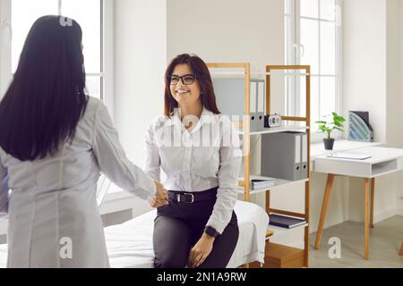 Happy female patient shaking hands with her doctor during appointment in medical office. Stock Photo