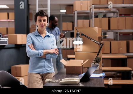 Warehouse manager standing in delivery department with arm crossed after finishing packing clients orders preparing packages for shipping. Diverse team working distribution center fulfillment company Stock Photo