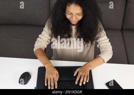 Cheerful black woman working on laptop computer at home. Overhead photo of BIPOC female in 30s doing distant work on notebook pc Stock Photo